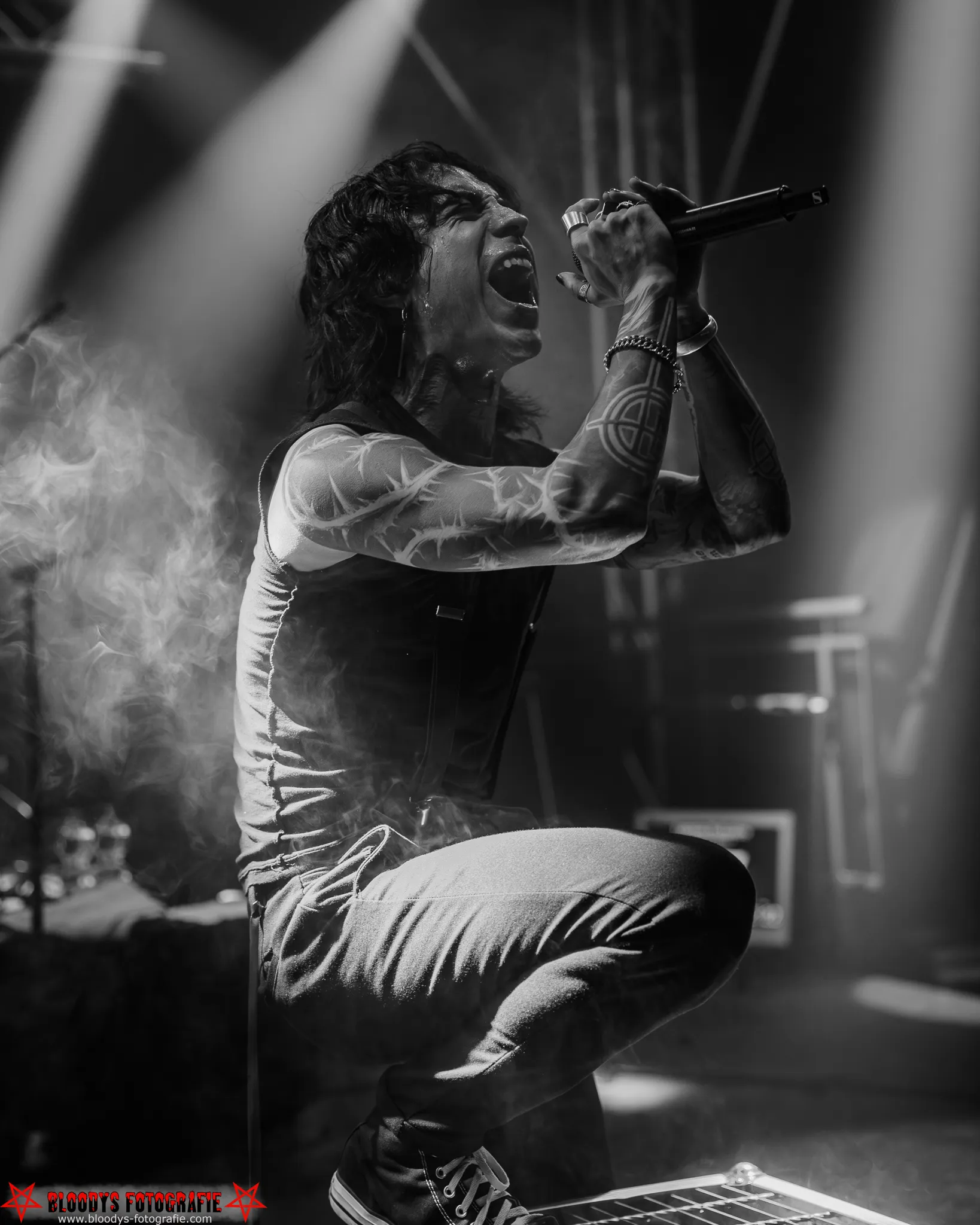 Black-and-white image of a tattooed singer passionately performing on stage holding a microphone, illuminated by dramatic stage lights and surrounded by smoke.