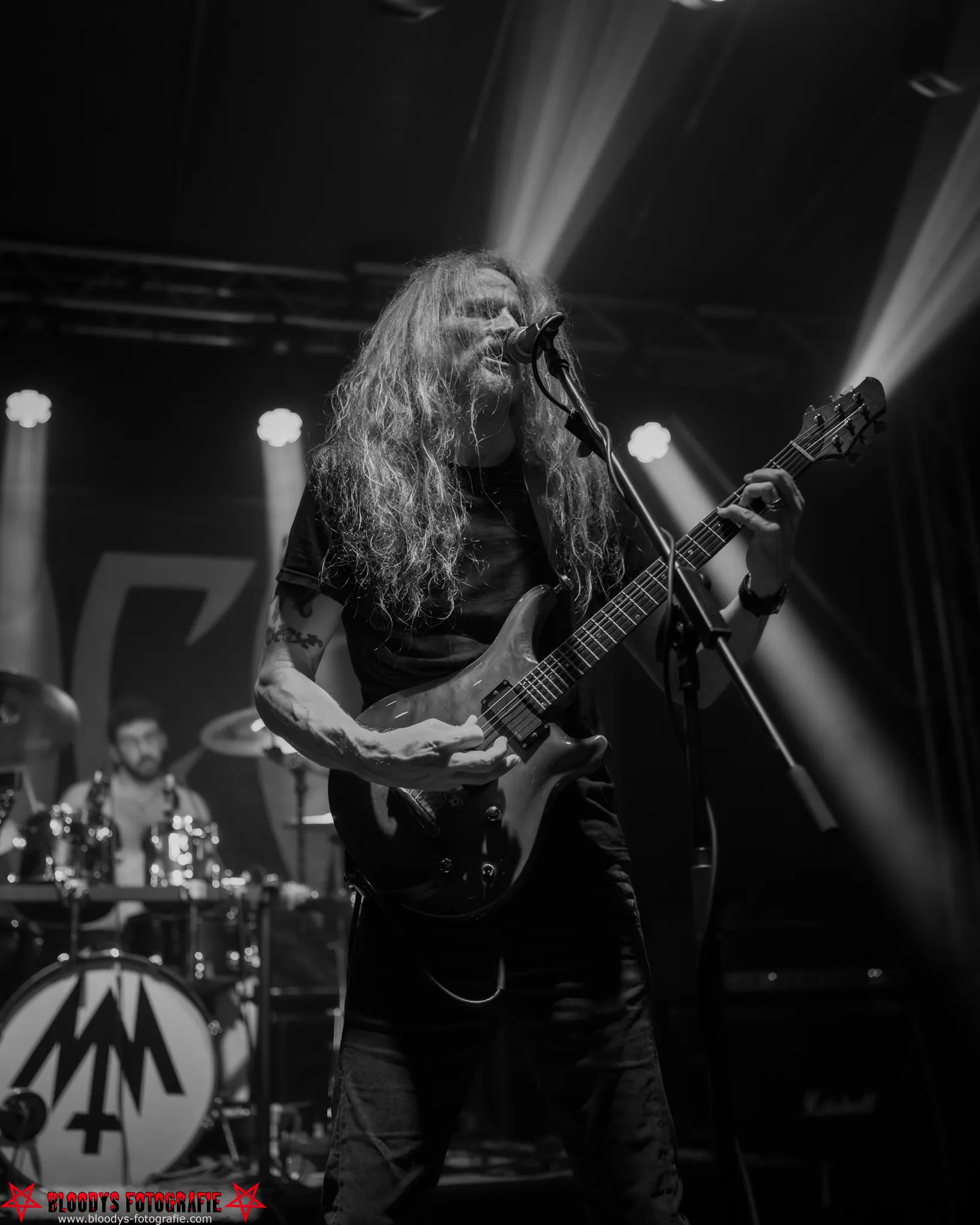 Long-haired guitarist singing into a microphone on stage with a drummer and drum set in the background, illuminated by stage lights.