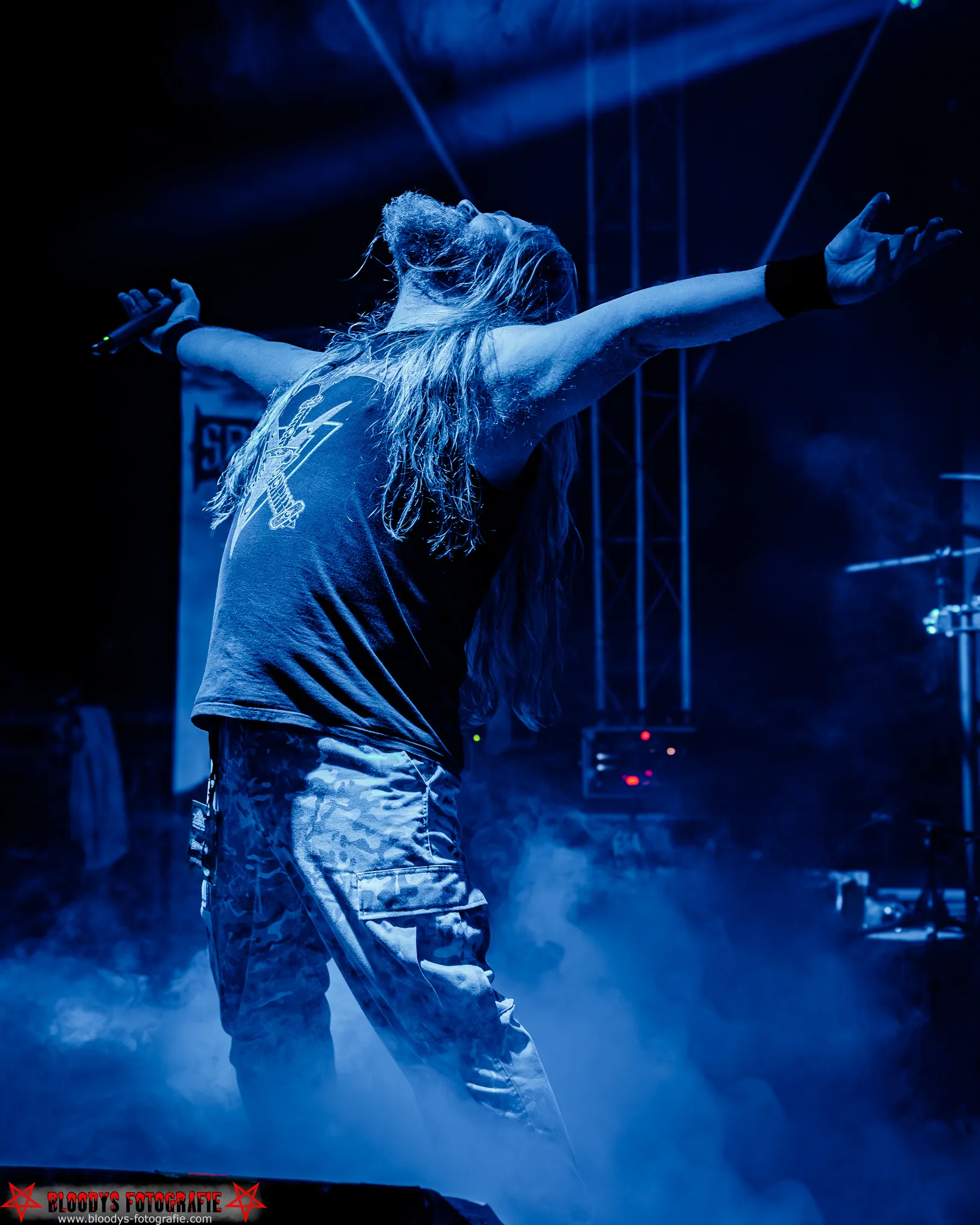 Long-haired male rock singer with arms outstretched, performing on stage with blue lighting and smoke.