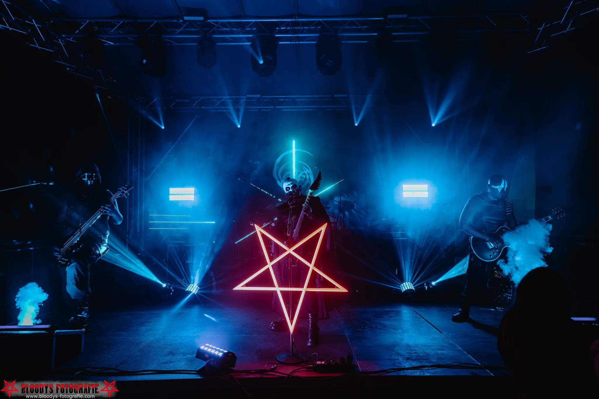 Metal band performing on stage with blue lighting and a neon pentagram in front of the lead singer wearing face paint.