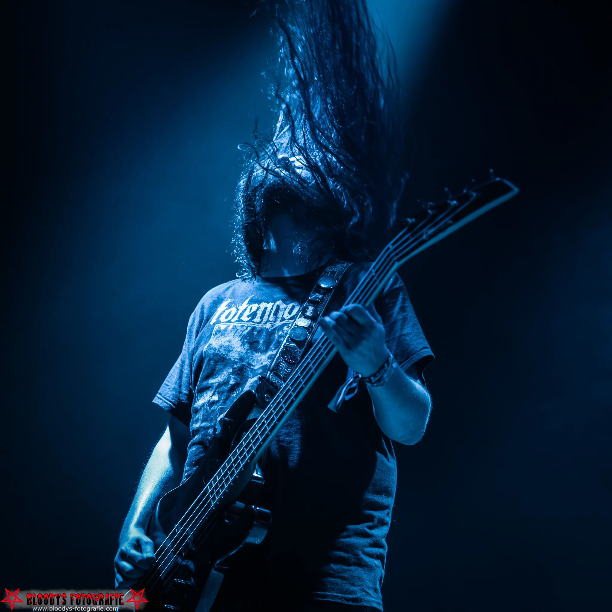 Musician with long hair playing bass guitar on stage under blue lighting.