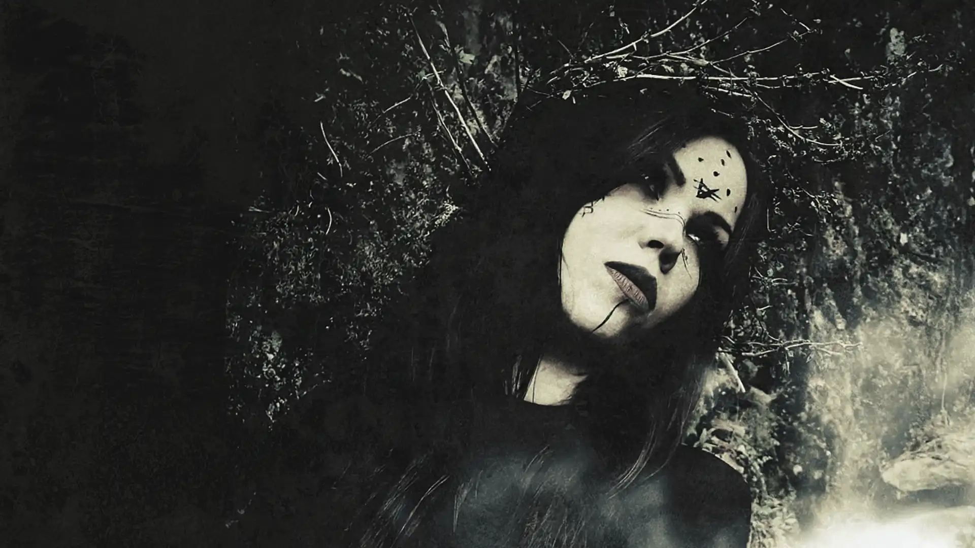 Woman with dark hair and black mystical face paint, adorned with a crown of twigs, against a dark, forested background. Band: Vermilia