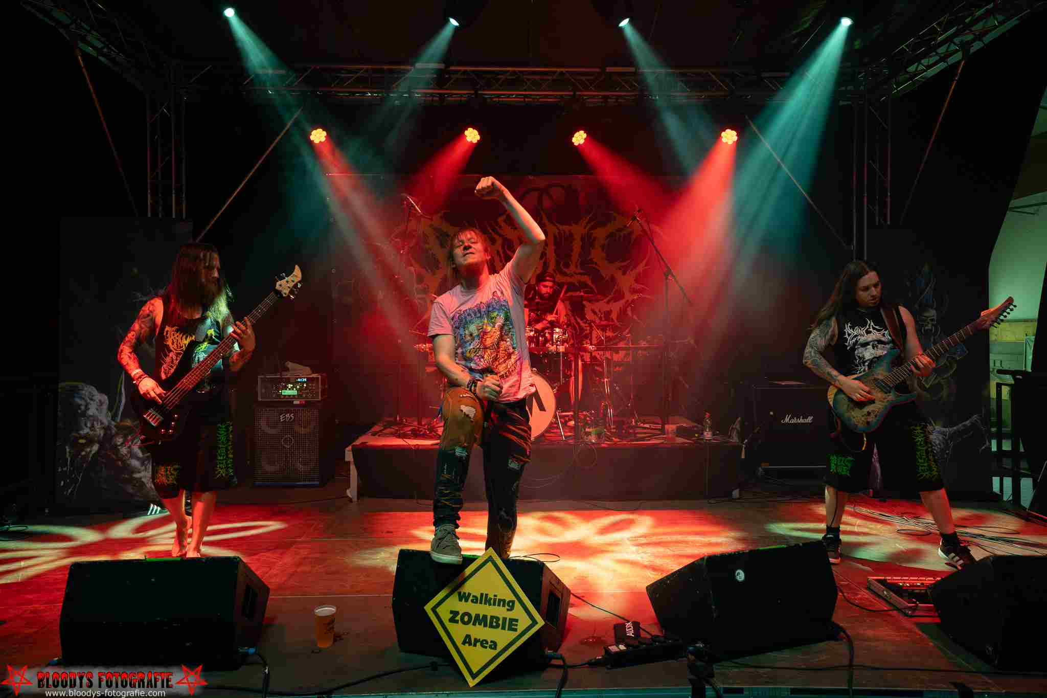 Metal band performing on stage with red and teal spotlights and a sign reading 'Walking ZOMBIE Area' in front.