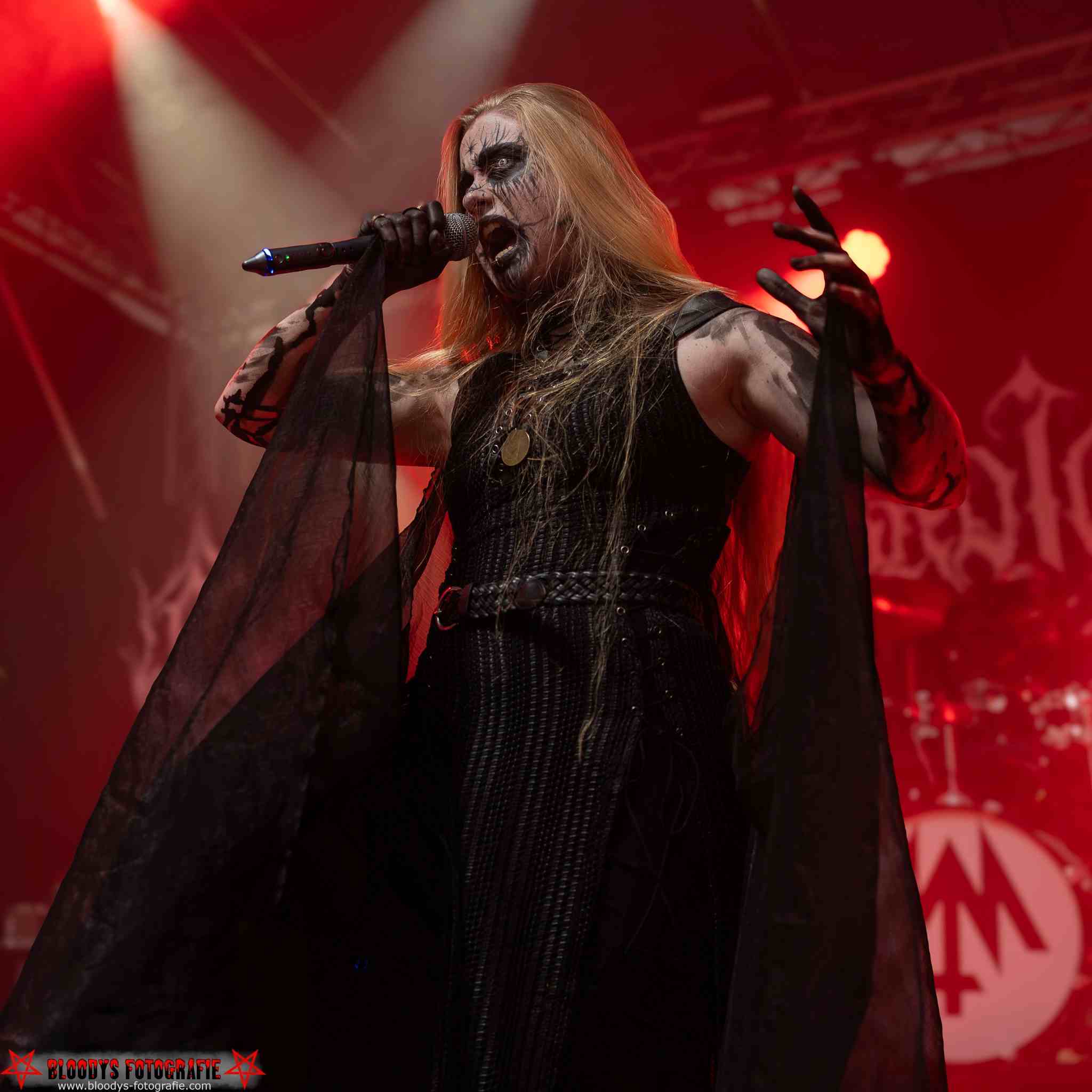 Heavy metal singer with corpse paint, long blonde hair, and black outfit passionately performing on stage with red lighting.
