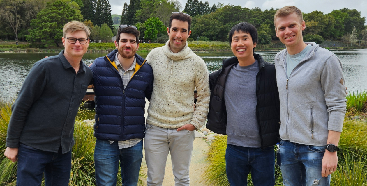 Five Opsin team members standing close together smiling in front of a lake with trees and greenery in the background.