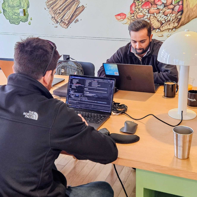 Two Opsin team members working on laptops at a wooden table in a casual office space with colorful wall art of food items. The table holds computer accessories, mugs, and a modern white desk lamp.