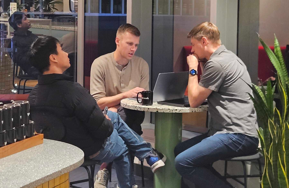 Three Opsin founders sitting around a small round table with a laptop, engaged in a discussion in a modern indoor setting with large windows and plants nearby.