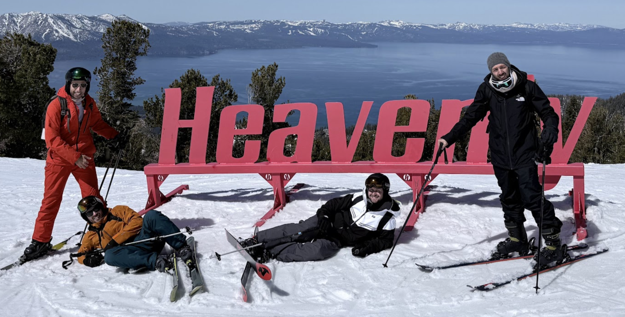 Four Opsin team members in winter gear posing on snow in front of a large pink 'Heavenly' sign with a lake and snow-covered mountains in the background under a clear blue sky.
