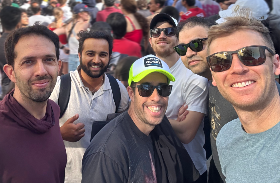 Group selfie of six Opsin team members smiling outdoors in a crowded area, some wearing sunglasses and casual clothing.