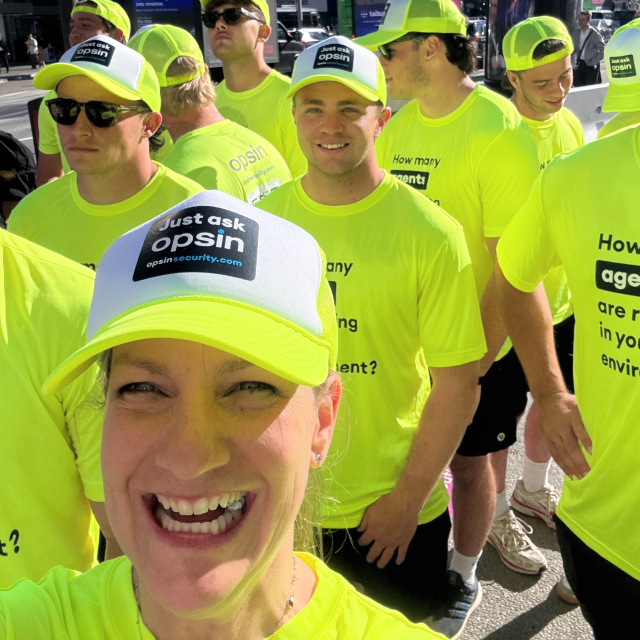 Opsin squad at an event wearing bright yellow shirts and matching caps that say 'Just ask Opsin' standing outdoors. The shirts have the text 'How many agents are running in your environment?' printed on them.