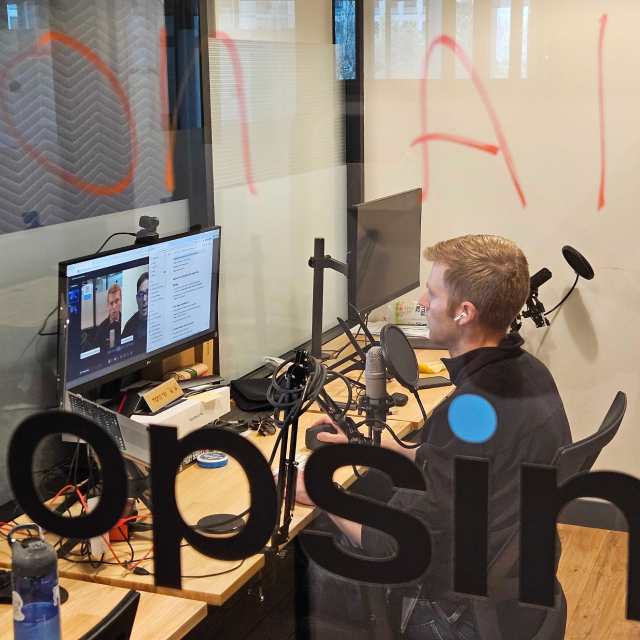 Opsin founder Oz Wasserman sits at a desk in an office booth, looking at a desktop monitor displaying a video call, with a microphone and audio equipment on the desk.