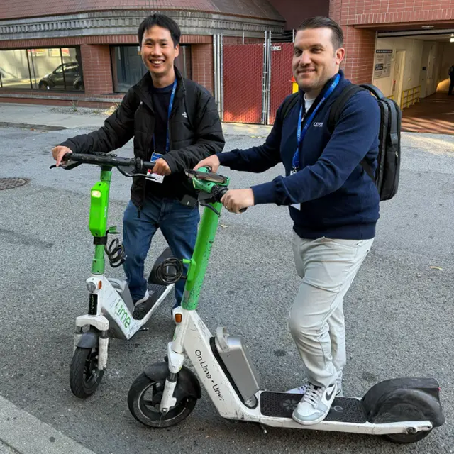 Two Opsin team members smiling and posing with electric scooters on a city street; one man is standing on the scooter, the other is holding the handlebar while standing beside it.