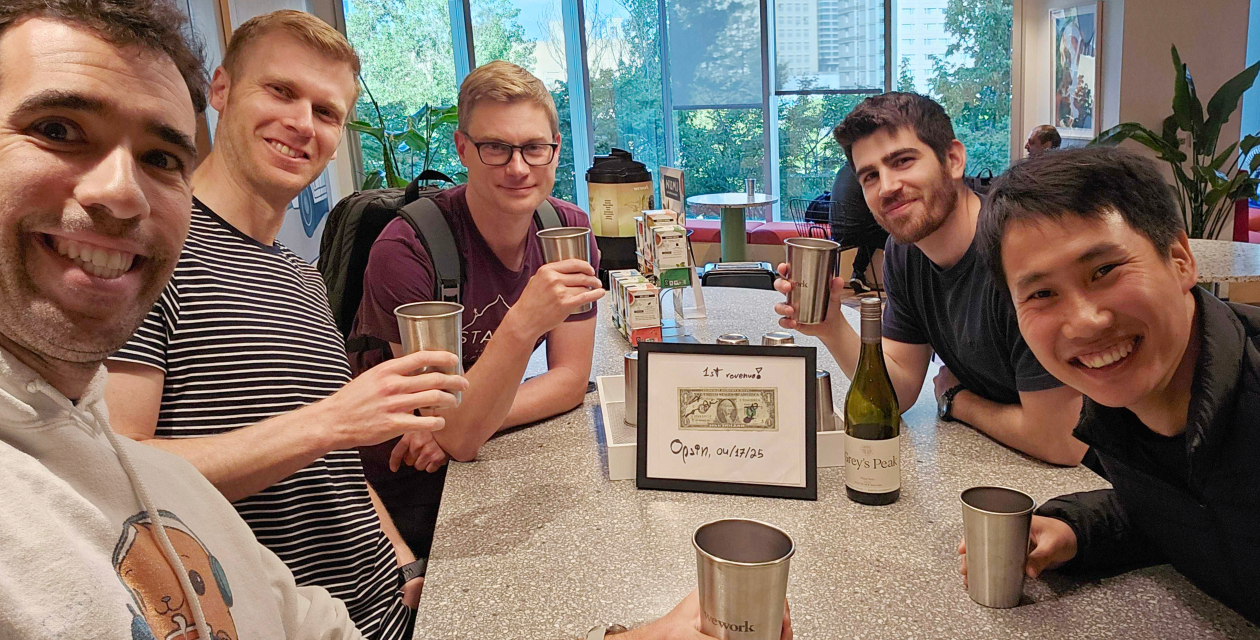 Five Opsin team members sitting around a table smiling and holding metal cups for a toast, in a bright office lounge with large windows and green plants outside.