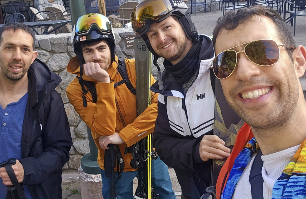 Four Opsin team members dressed in winter and ski gear smiling outdoors next to a stone wall; one wears sunglasses and colorful jacket, another has a ski helmet and goggles, holding skis, with outdoor chairs and tables in the background.