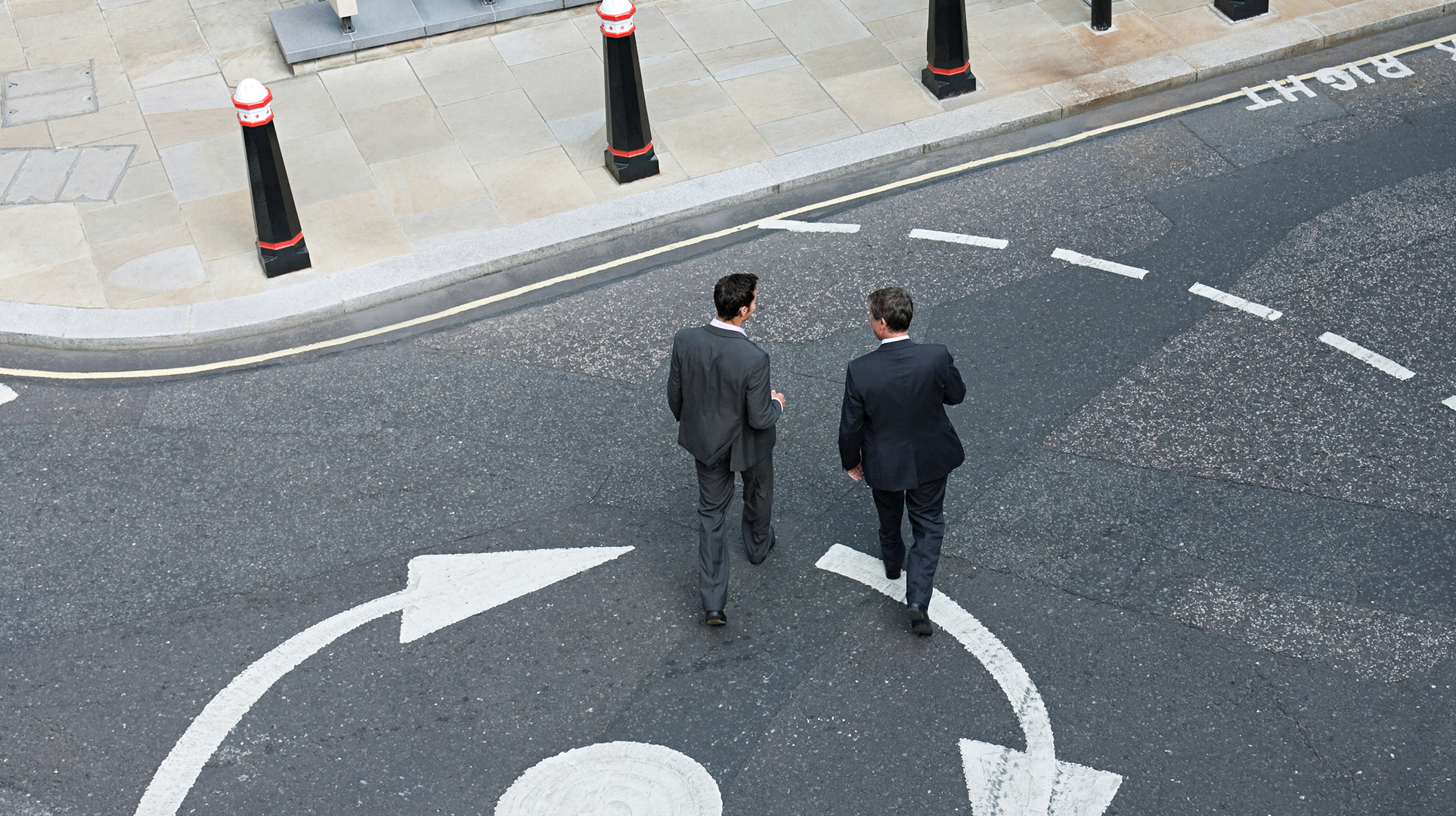 Two business professionals walking across a road with directional arrow markings.