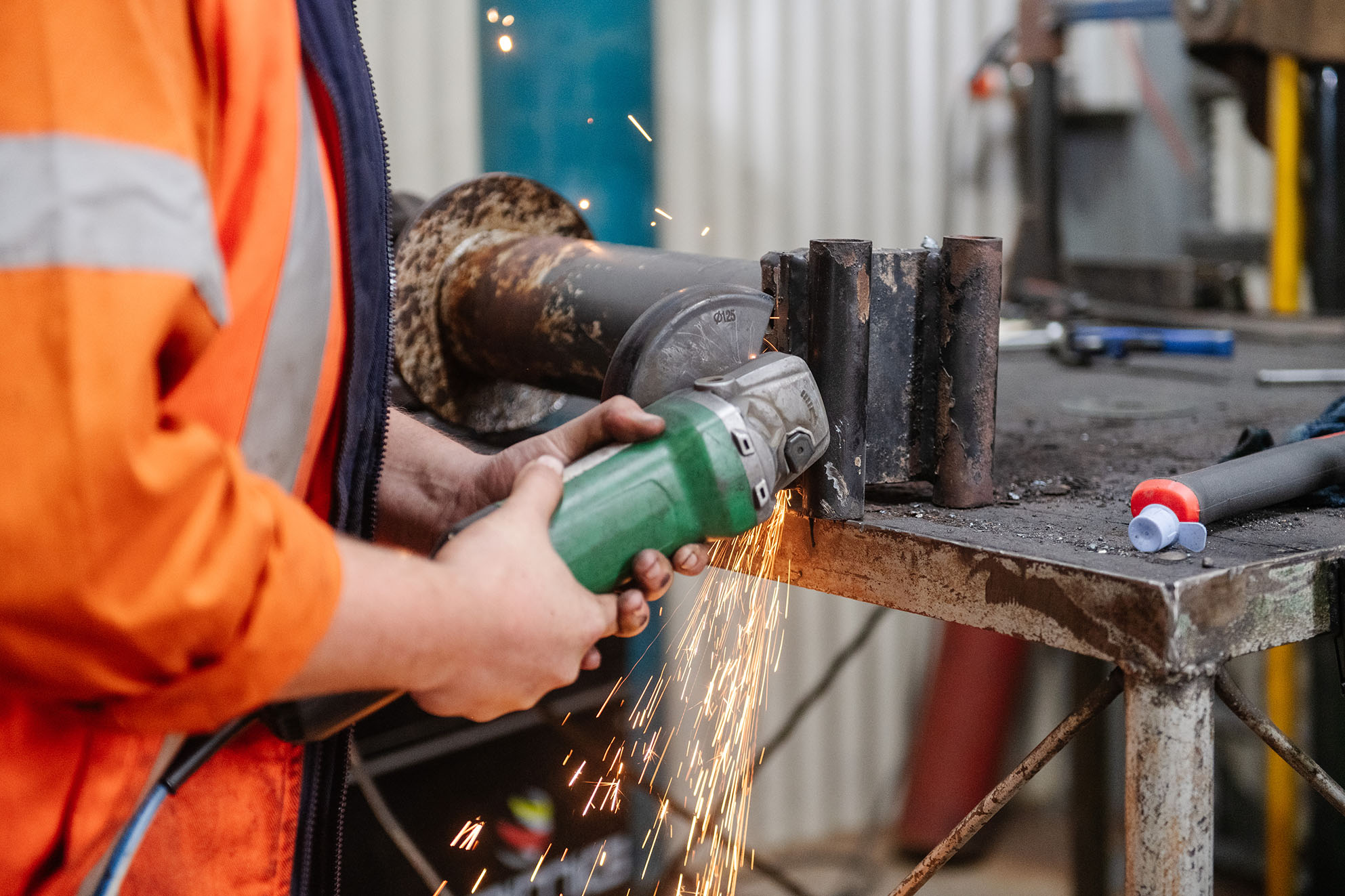 Worker in an orange high-visibility jacket using an angle grinder on a metal pipe, producing sparks in an industrial workshop.