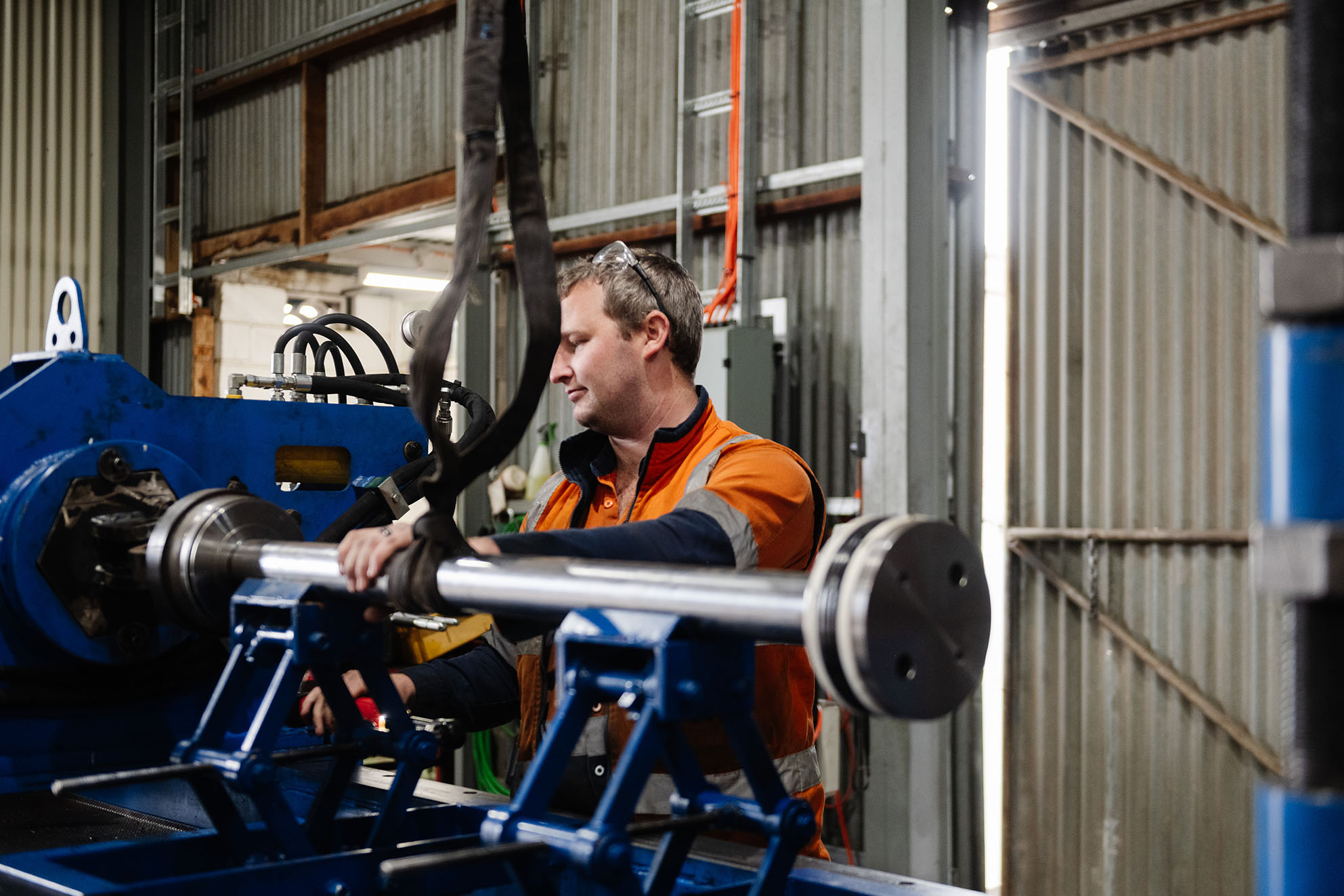 Worker in high-visibility clothing operating machinery and inspecting a metal shaft in an industrial workshop setting.