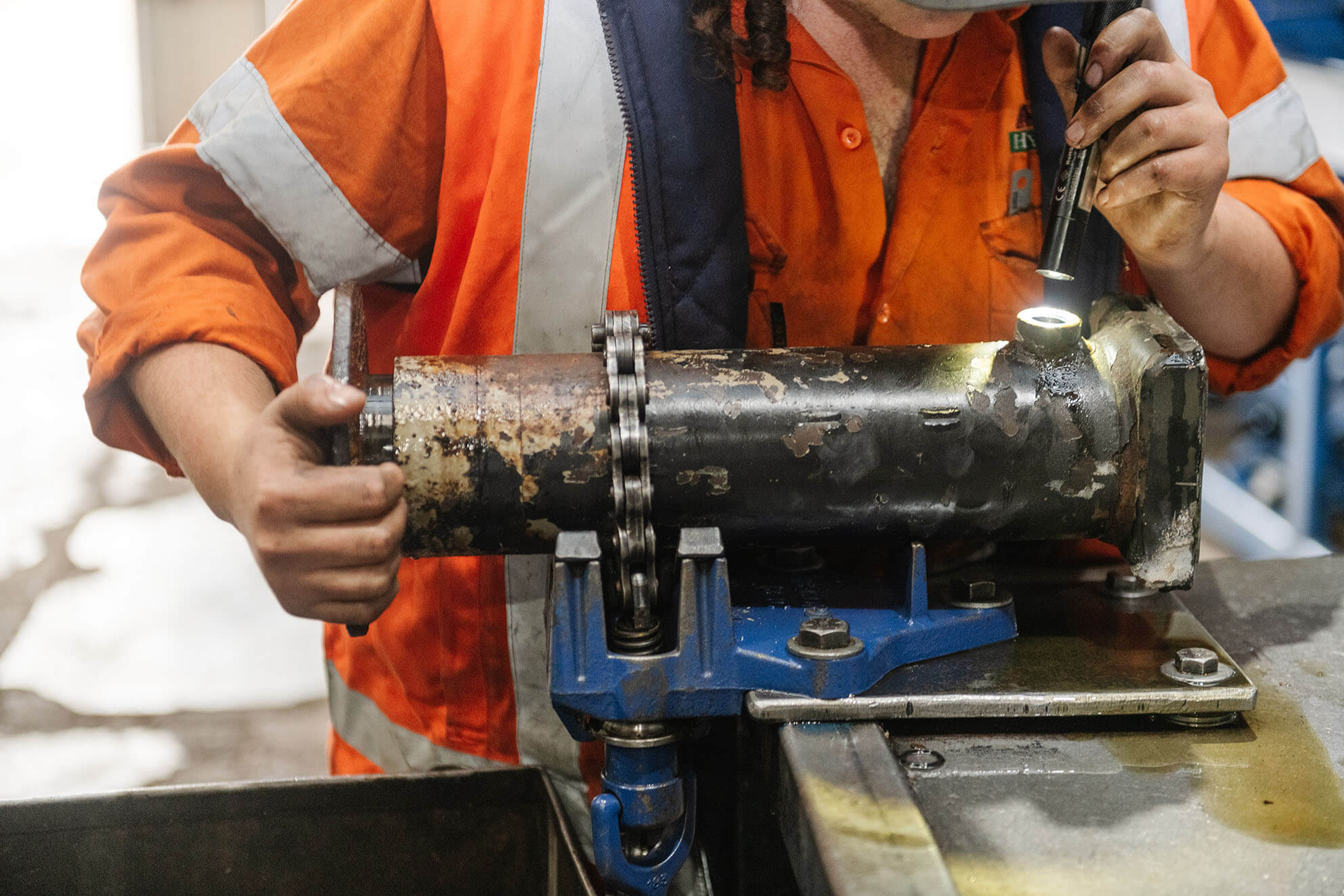 Worker in high-visibility clothing inspecting a metal component with a flashlight, secured in a vice on a workbench in an industrial workshop.