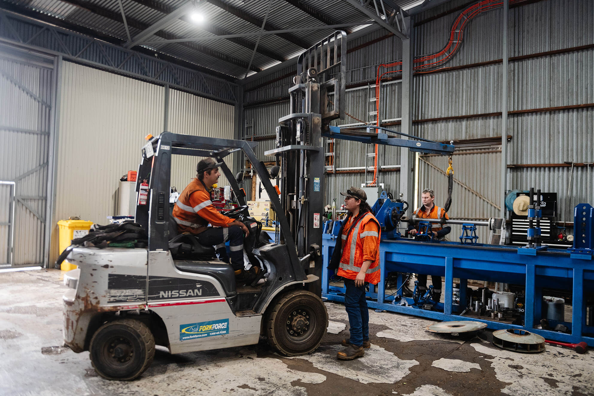 Two workers in safety vests operating forklift inside workshop.