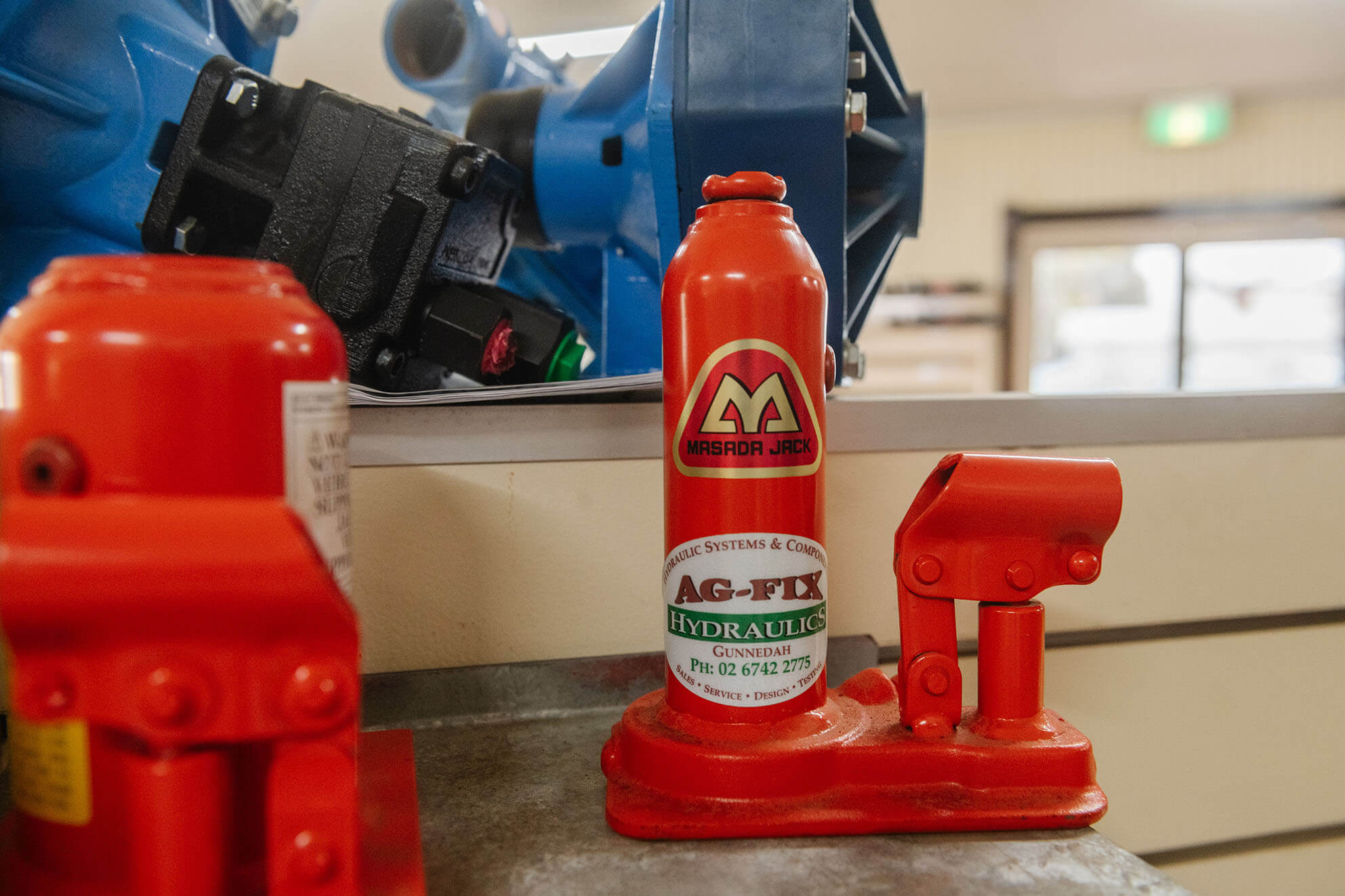 Red hydraulic bottle jack with Ag-Fix branding on workshop counter.