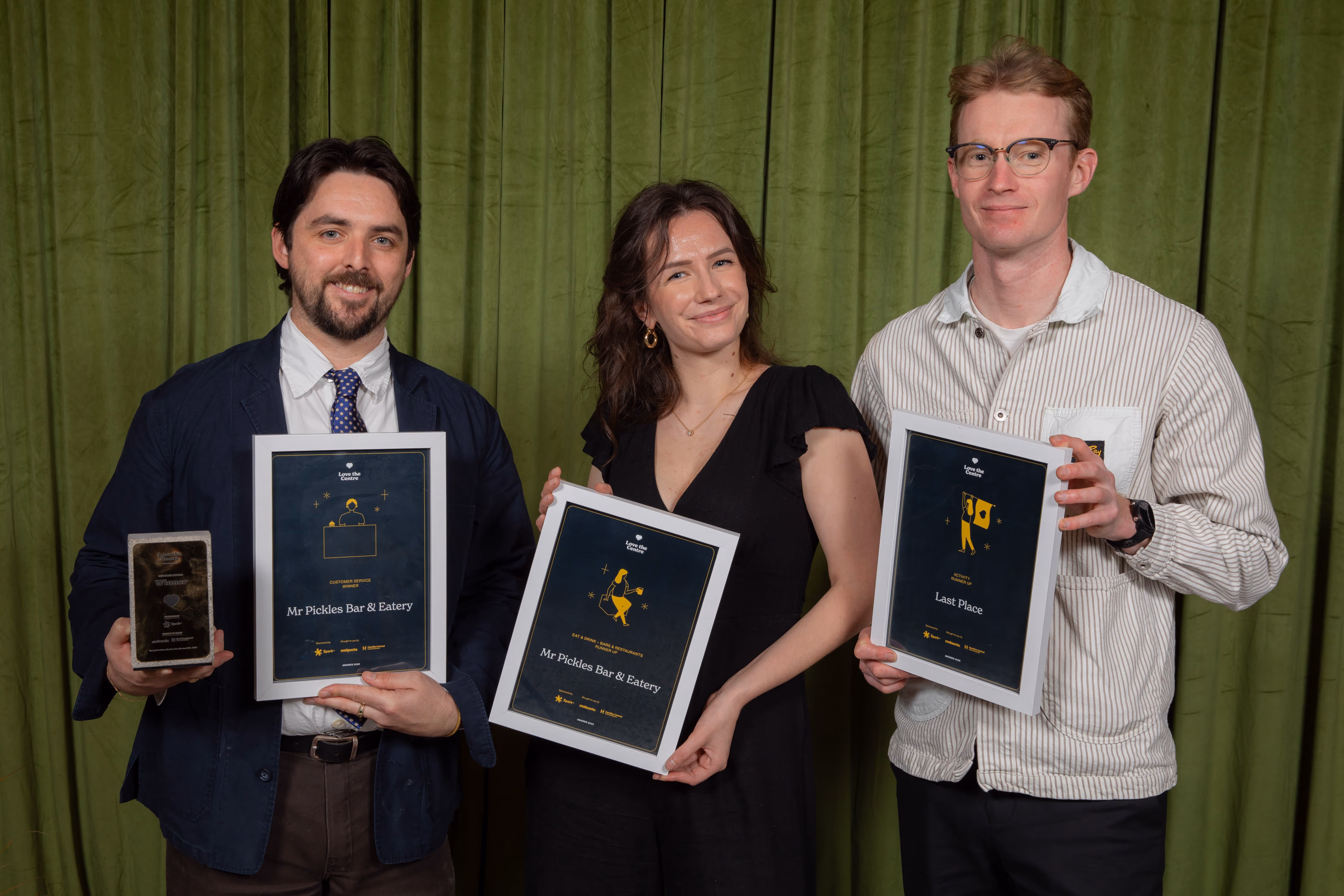 Three people smiling and holding framed awards against a green curtain backdrop.