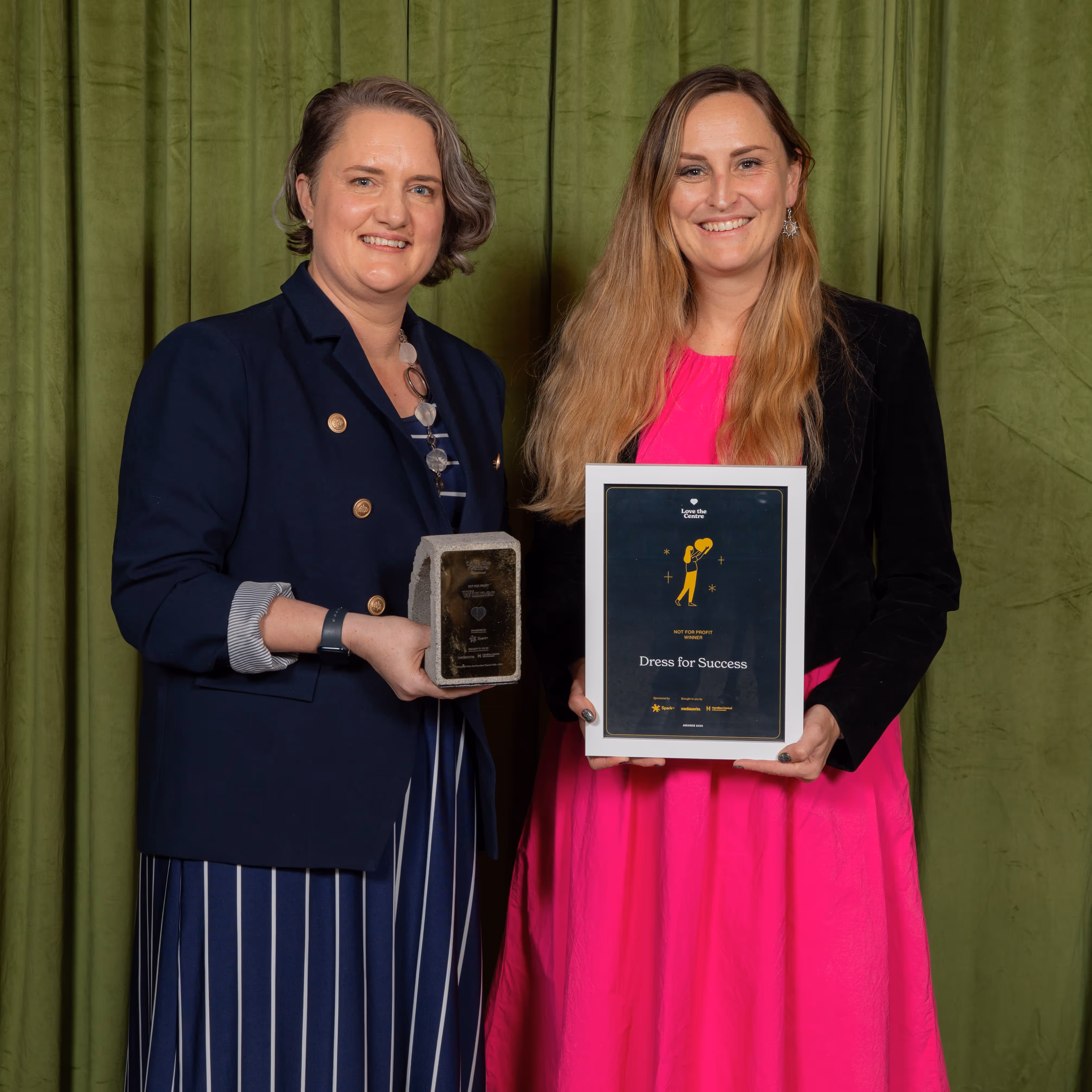 Two women smiling and holding awards against a green curtain backdrop.