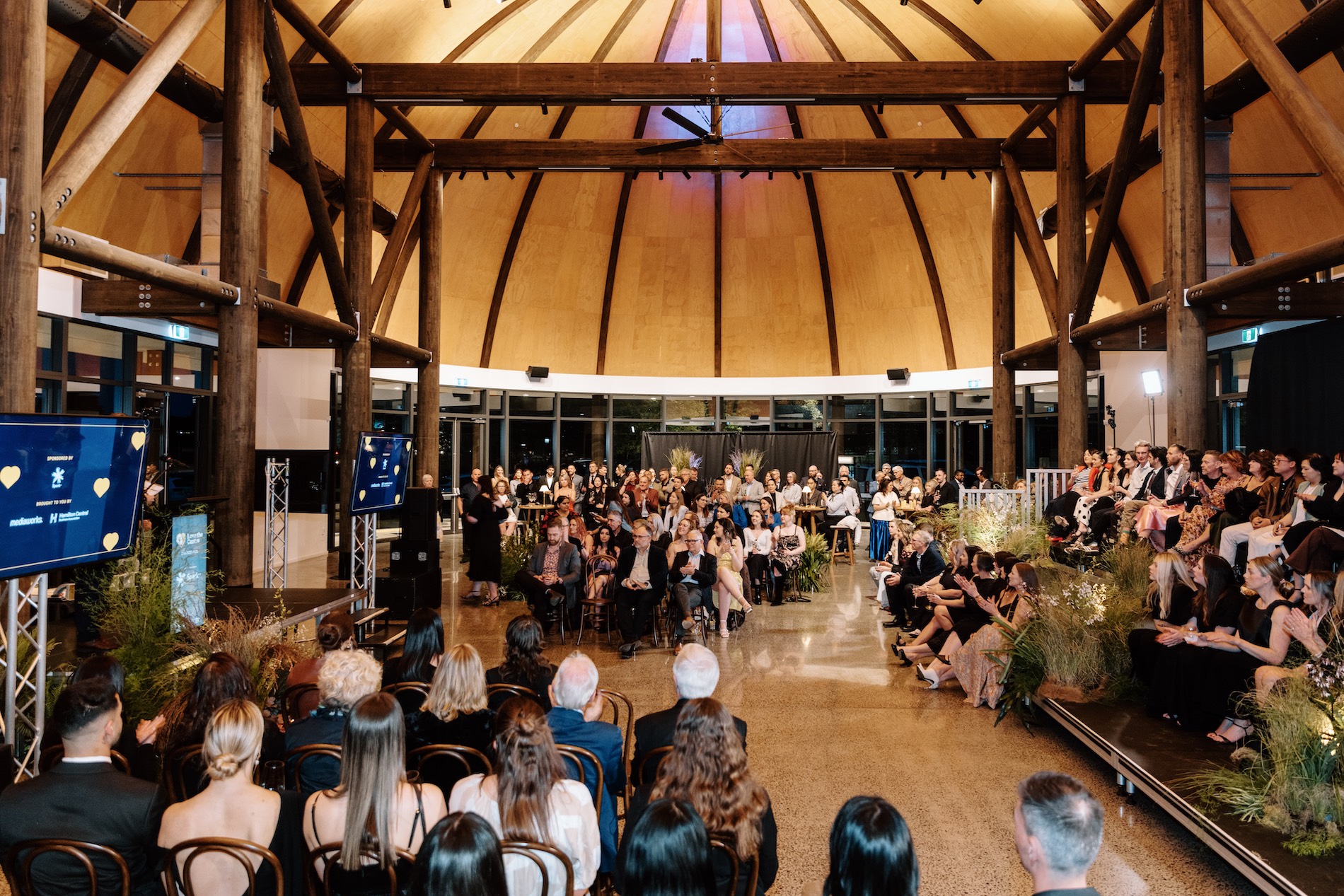 Audience seated in a circular venue with exposed timber beams watching a presentation or ceremony.