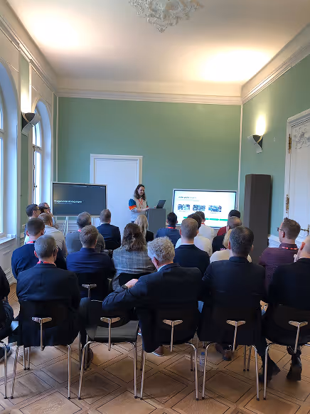 Person hält eine Präsentation vor einem Publikum in einem Raum mit grünen Wänden und großen Fenstern.