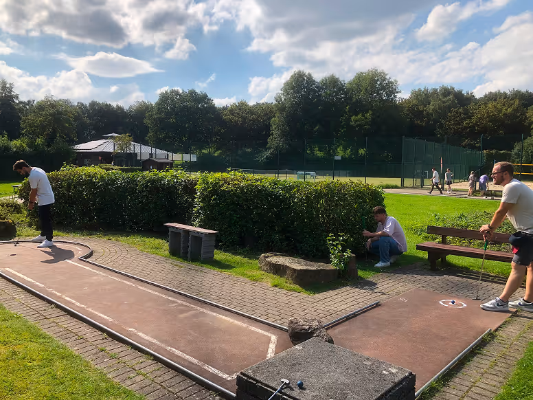 Menschen spielen Minigolf auf einem sonnigen Außenplatz mit grünen Büschen und Bäumen im Hintergrund.