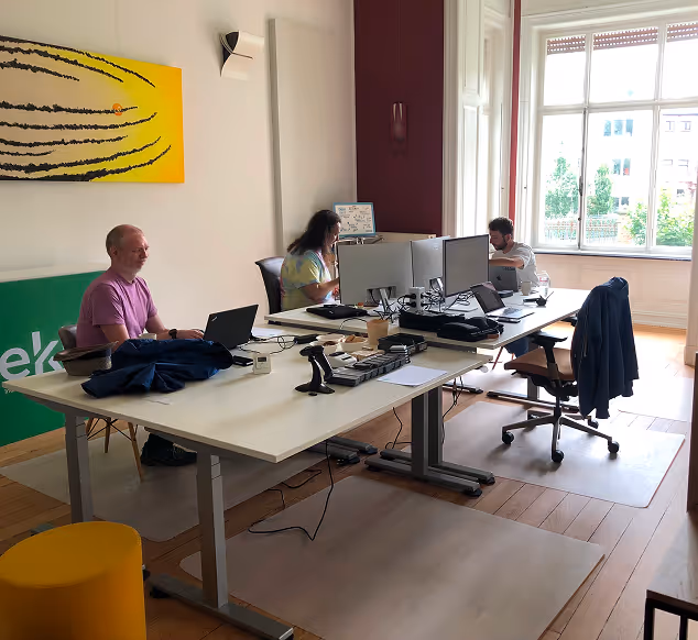 Drei Personen arbeiten an Computern an zwei großen weißen Tischen in einem hellen Büro mit großem Fenster.