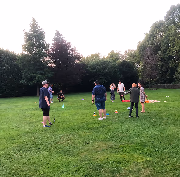 Gruppe von Menschen spielt und unterhält sich auf einer grünen Wiese im Freien, umgeben von Bäumen.