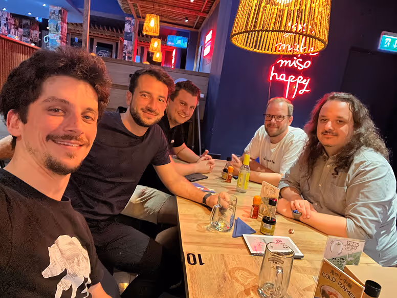 Fünf Männer sitzen an einem Holztisch in einem gemütlichen Restaurant mit Neonlicht und lächeln in die Kamera.