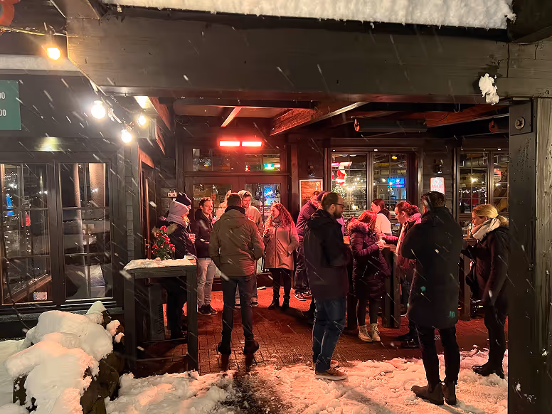 Gruppe von Menschen in Winterkleidung, die sich nachts unter einem überdachten Bereich mit Schneeboden und Beleuchtung unterhalten.