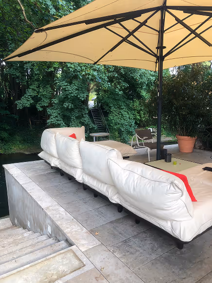 Große weiße Außenlounge mit mehreren Kissen unter einem großen Sonnenschirm neben einer Treppe, umgeben von grünen Bäumen.