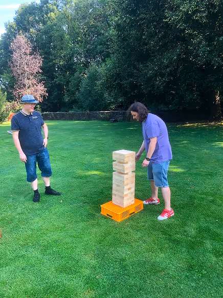 Zwei Männer spielen draußen auf einer Wiese ein übergroßes Jenga-Spiel aus Holzwürfeln auf einer orangen Kiste.