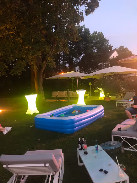 Abendliche Gartenparty mit aufblasbarem Pool, leuchtenden Stehtischen, Sonnenschirmen und Gartenmöbeln auf Rasen.