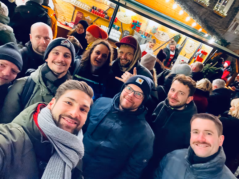 Gruppe von Menschen in Winterkleidung, die bei einem belebten Marktstand lächeln und sich für ein Selfie versammeln.