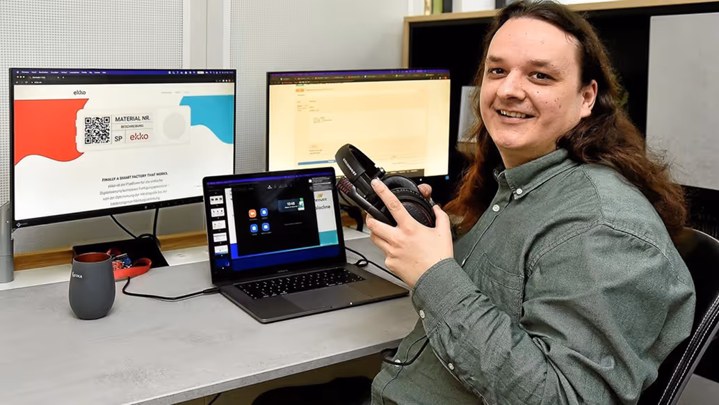 Mann mit langem braunem Haar und grünem Hemd hält Kopfhörer und sitzt an einem Schreibtisch mit drei Bildschirmen.