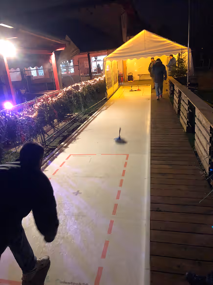 Menschen spielen Shuffleboard auf einer beleuchteten Bahn neben einem Zelt bei Nacht.