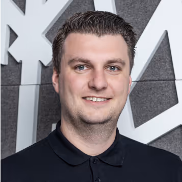 Porträt eines lächelnden Mannes mit kurzen braunen Haaren vor einer grauen Wand mit weißem abstraktem Muster.