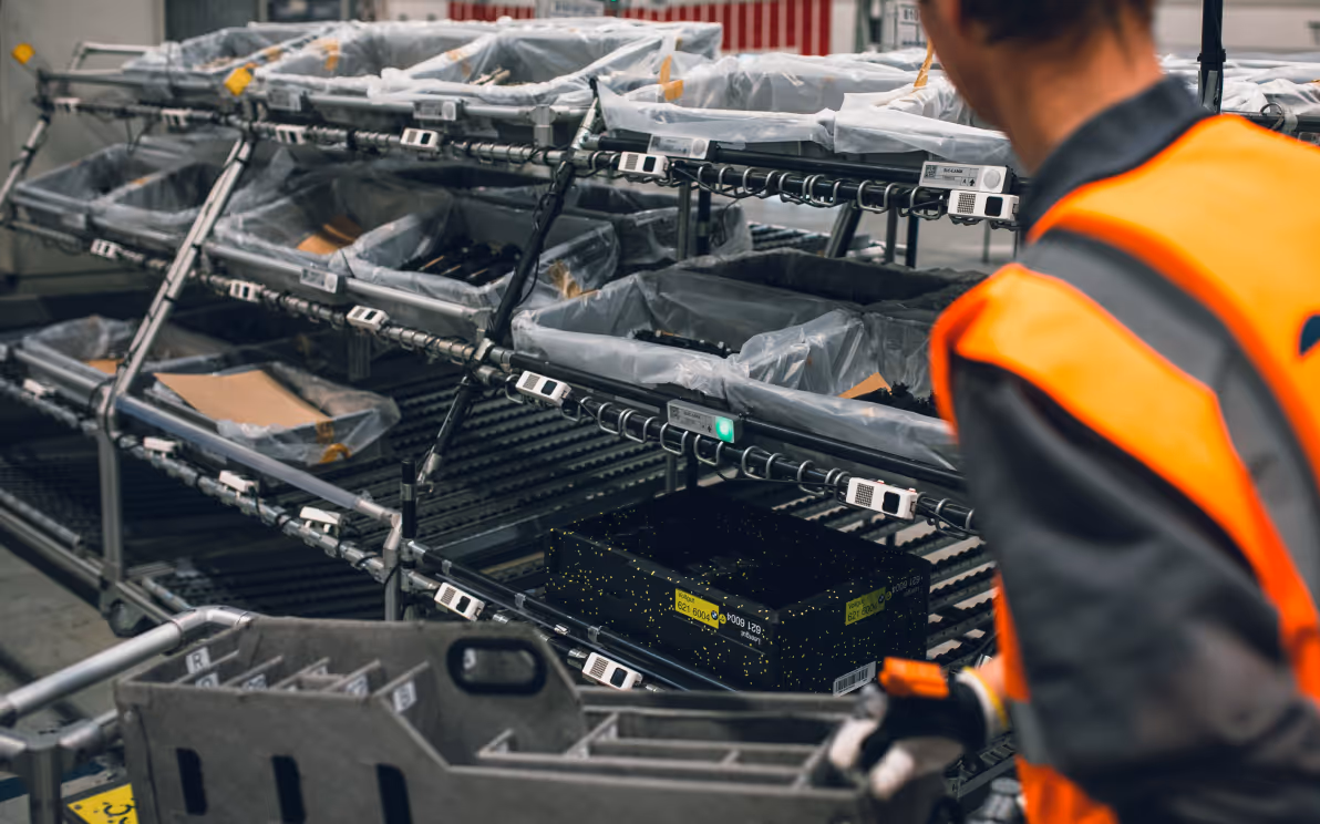 Mitarbeiter in Warnweste arbeitet in Lagerumgebung mit Pick-by-Light ausgestatten Regalen voller aufgeordneter Behälter und einem Rollwagen und Handscanner.