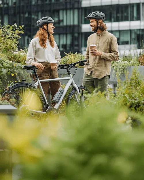 Zwei Personen mit Fahrradhelmen stehen mit einem Fahrrad im Grünen und halten Kaffeebecher, im Gespräch vor einem modernen Bürogebäude.
