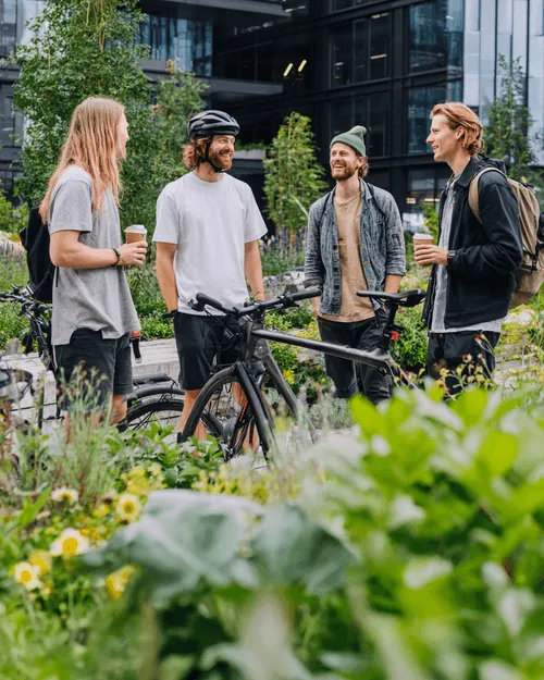Vier junge Männer unterhalten sich draußen um ein schwarzes Fahrrad, umgeben von grünen Pflanzen und moderner Architektur.