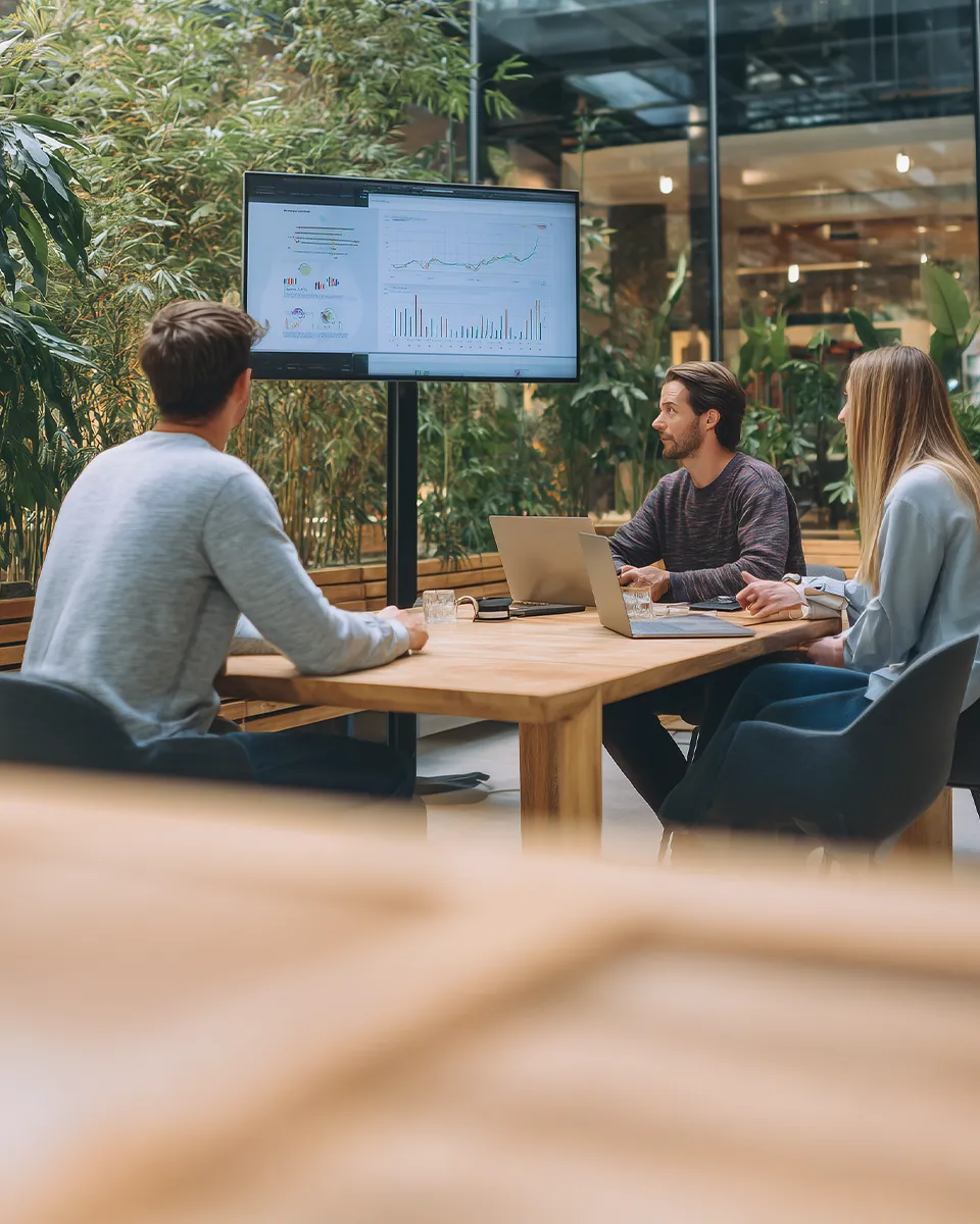Drei Personen in einem Meetingraum beobachten eine Bildschirmpräsentation mit Diagrammen und Diagrammen.