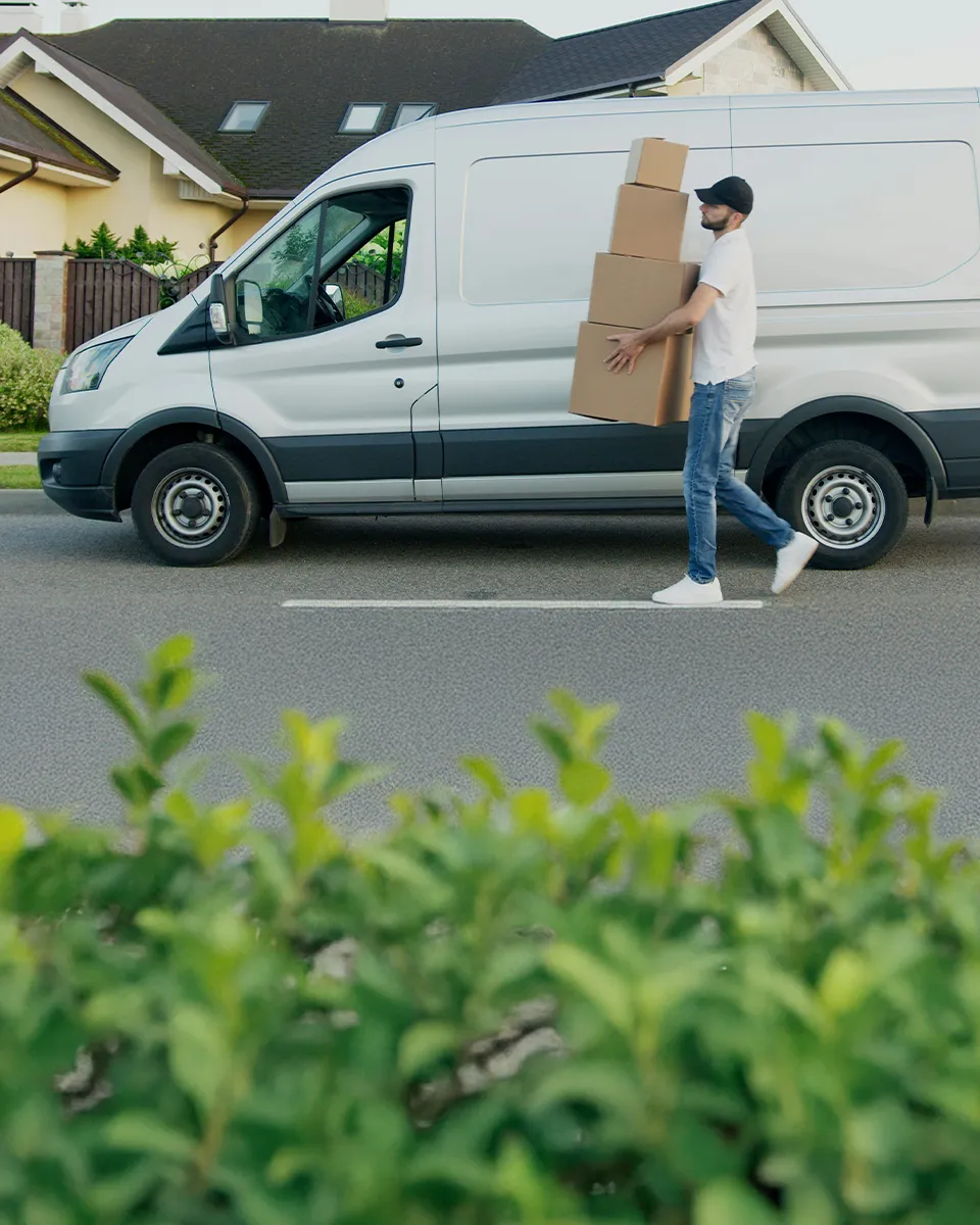 Ein Mann in Freizeitkleidung trägt mehrere Kartons vor einem geparkten weißen Lieferwagen auf einer Wohnstraße.