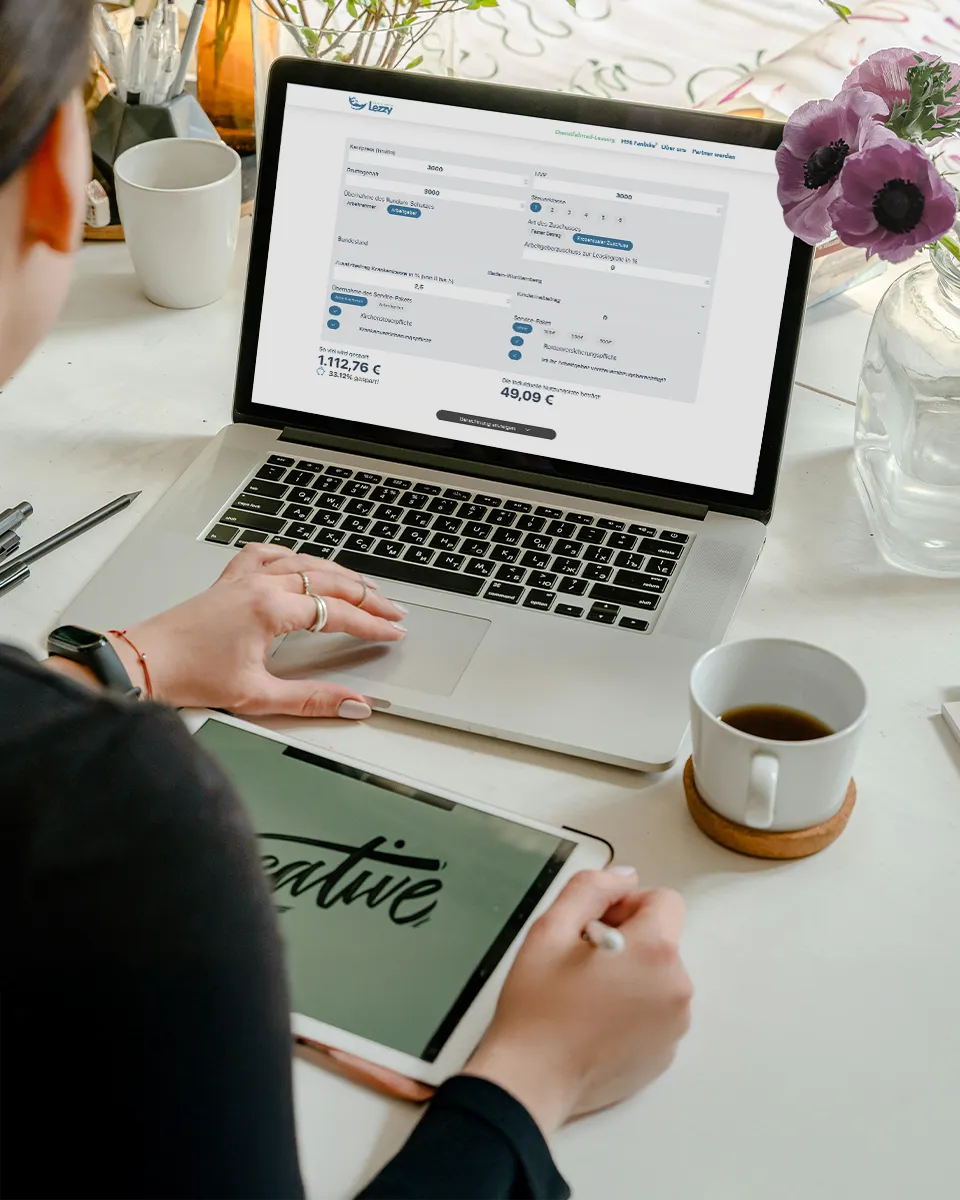Person nutzt Laptop mit Lezzy-Website und Tablet mit kreativem Schriftzug, neben Kaffeetasse und Blumen auf dem Tisch.