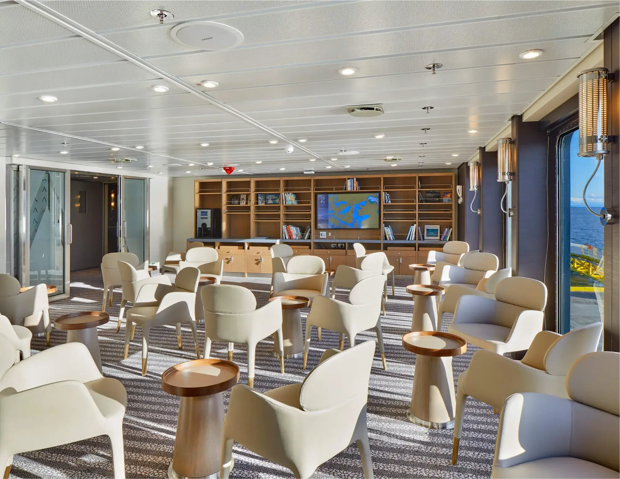 Bright lounge area with cream chairs and wooden round tables, bookshelves, and windows showing a view of the sea.