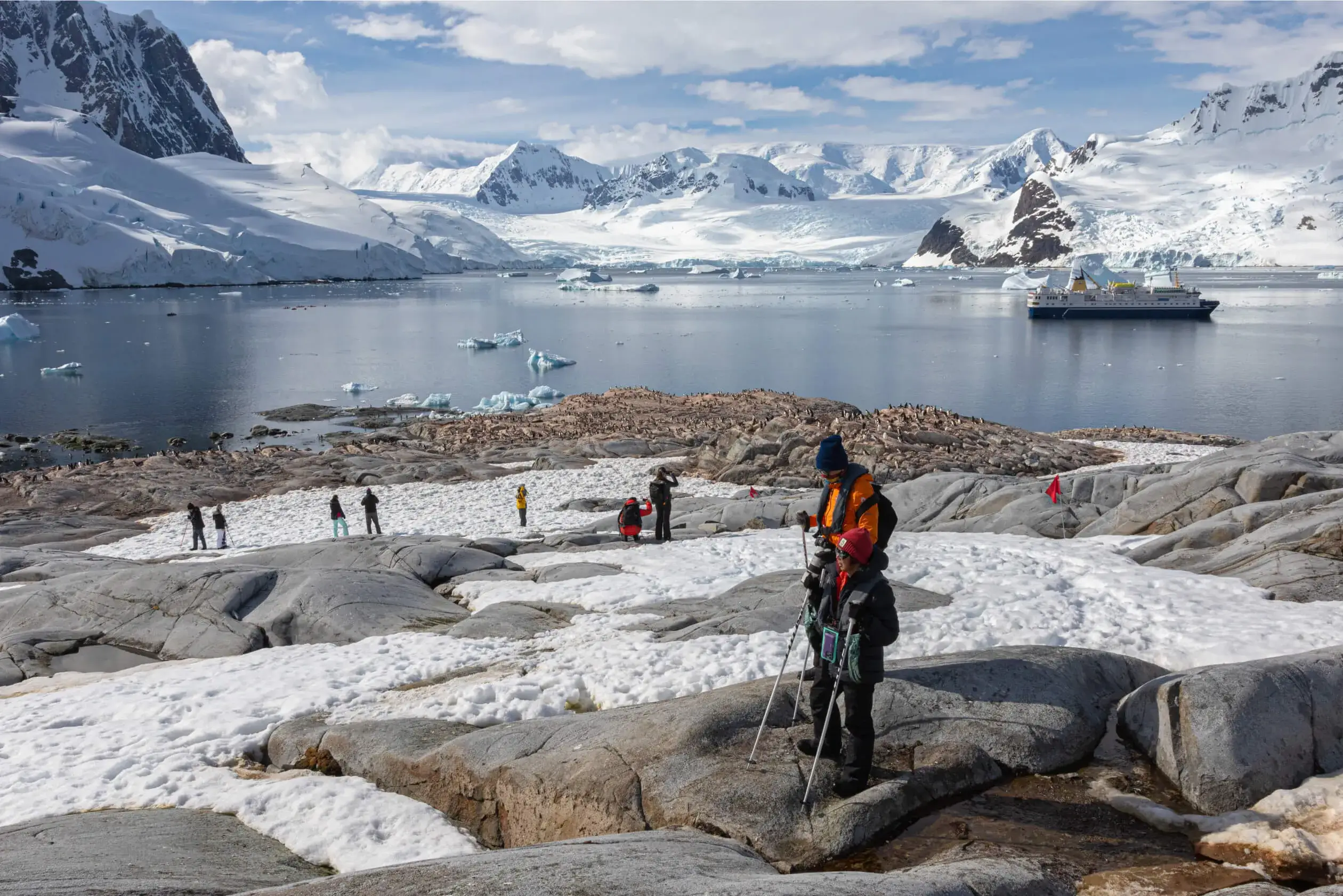 Group of tourists exploring a snowy rocky shore with penguins nearby, a ship in icy waters, and snow-covered mountains in the background.