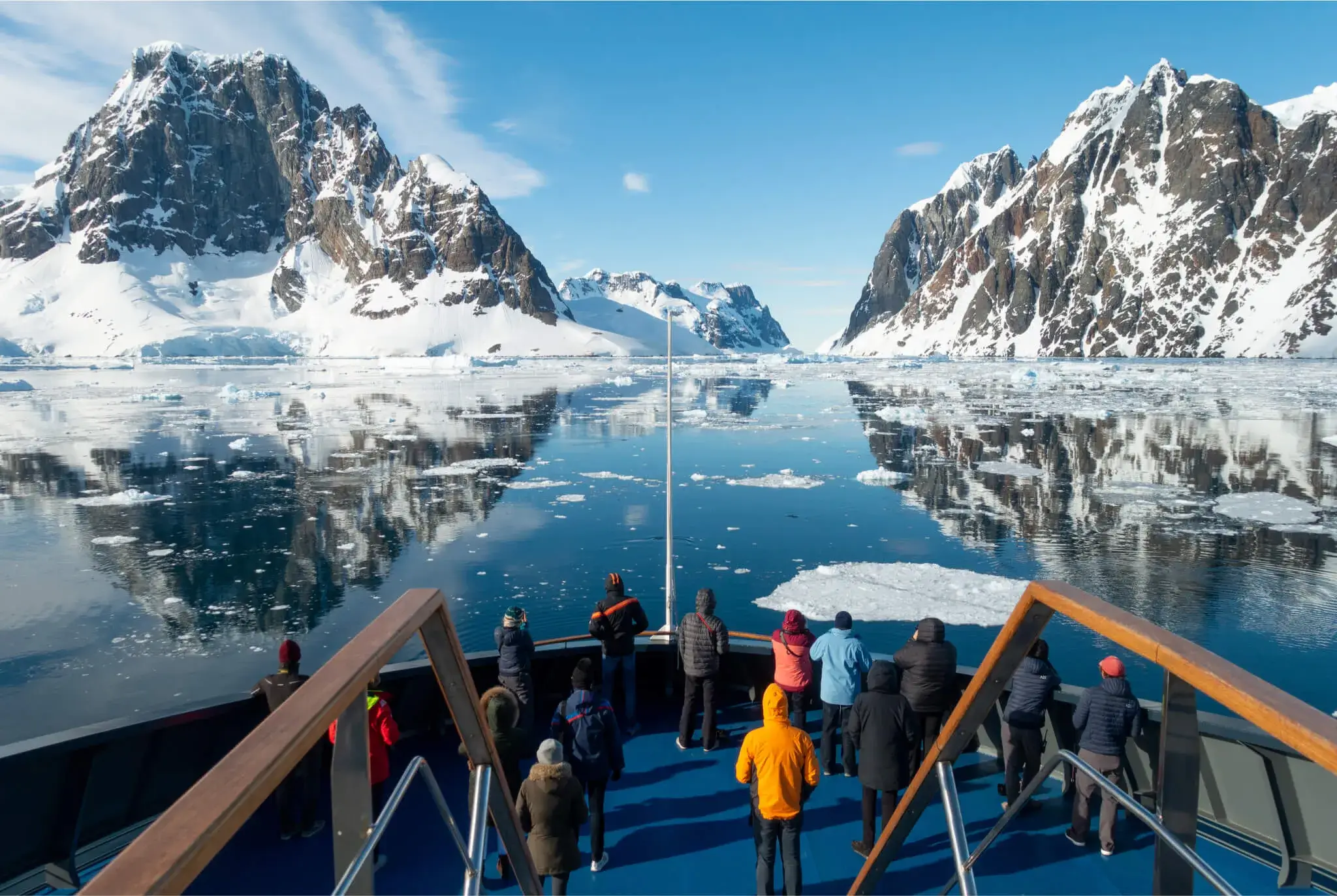 People on a boat observing snow-covered mountains and ice floating on calm water under a blue sky.