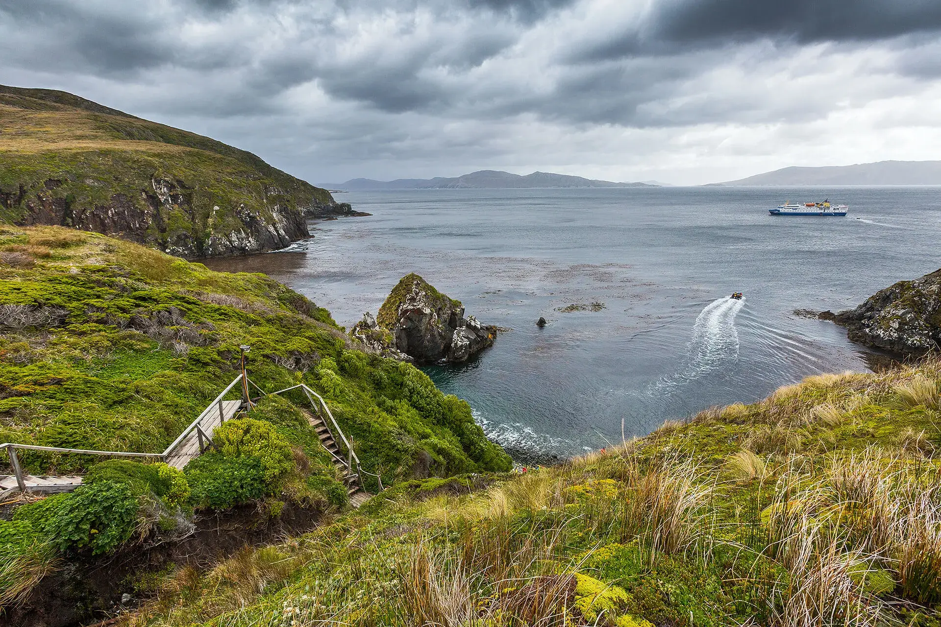 Coastal landscape with green cliffs, wooden stairs descending toward the water, a small boat leaving a wake, and a ferry on a cloudy day.